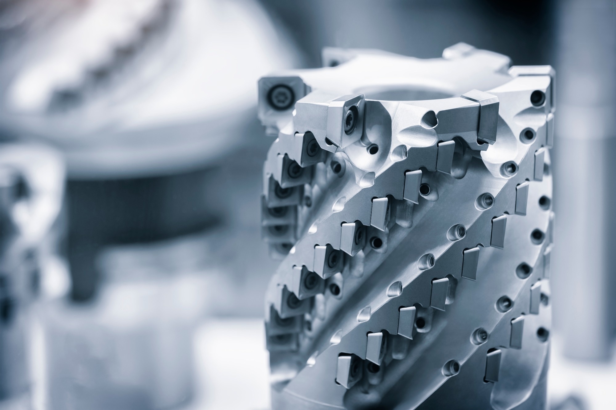 Close-up image of a precision-engineered cutting tool with multiple tungsten carbide inserts, designed for industrial machining applications. The tool features a spiral structure with sharp, replaceable cutting edges, set against a blurred workshop background.