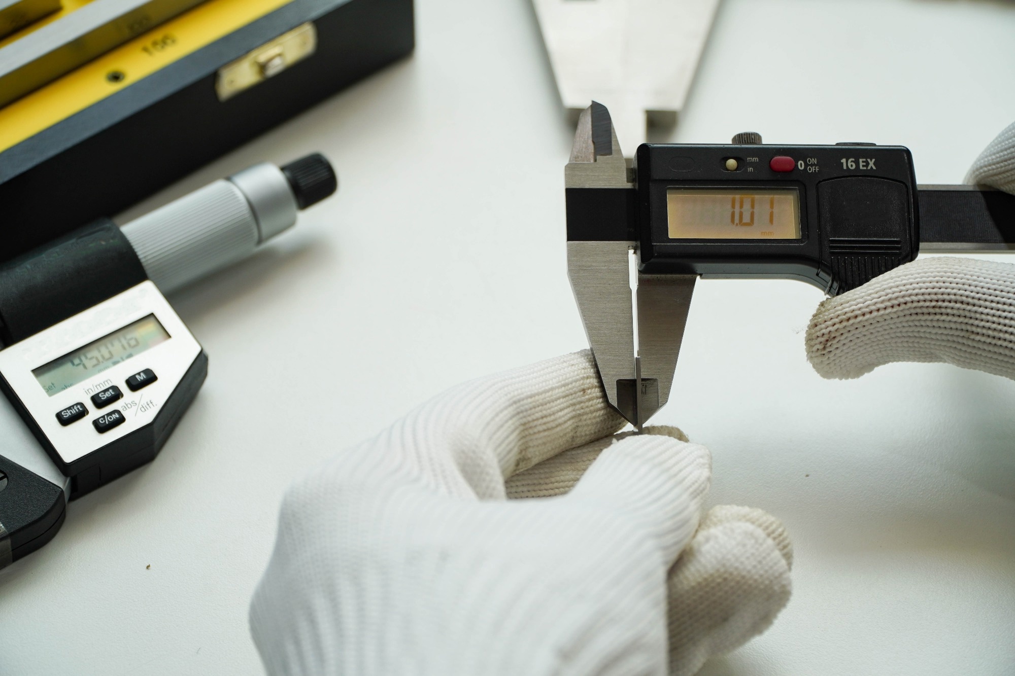 A close-up image of a person wearing white gloves using a digital vernier caliper to measure a small metal object. A digital micrometer, metal ruler, and precision tools are visible in the background, representing a metrology or quality control setup.