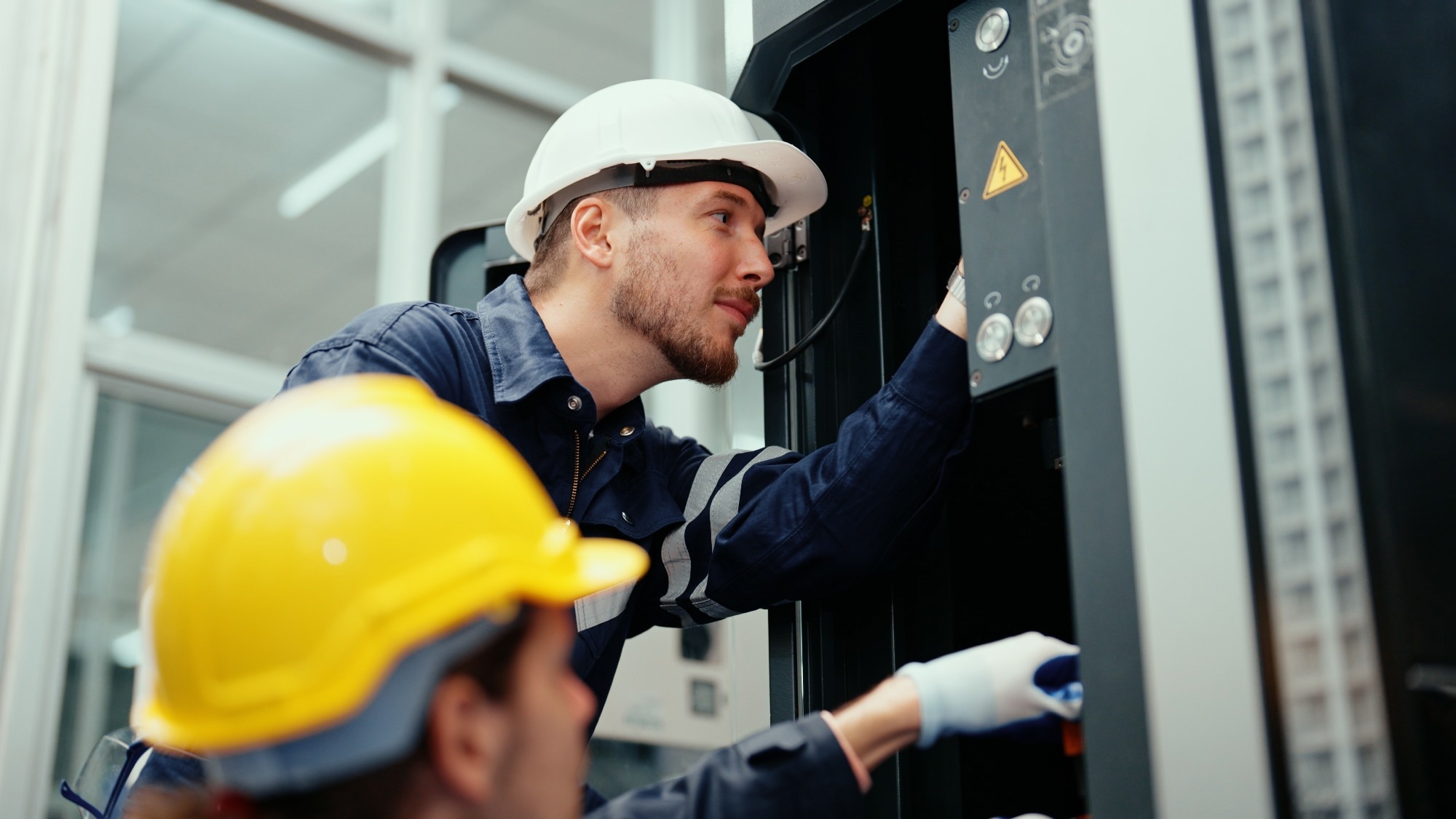 Two industrial workers wearing hard hats and safety gear inspect an electrical control panel in a modern facility.