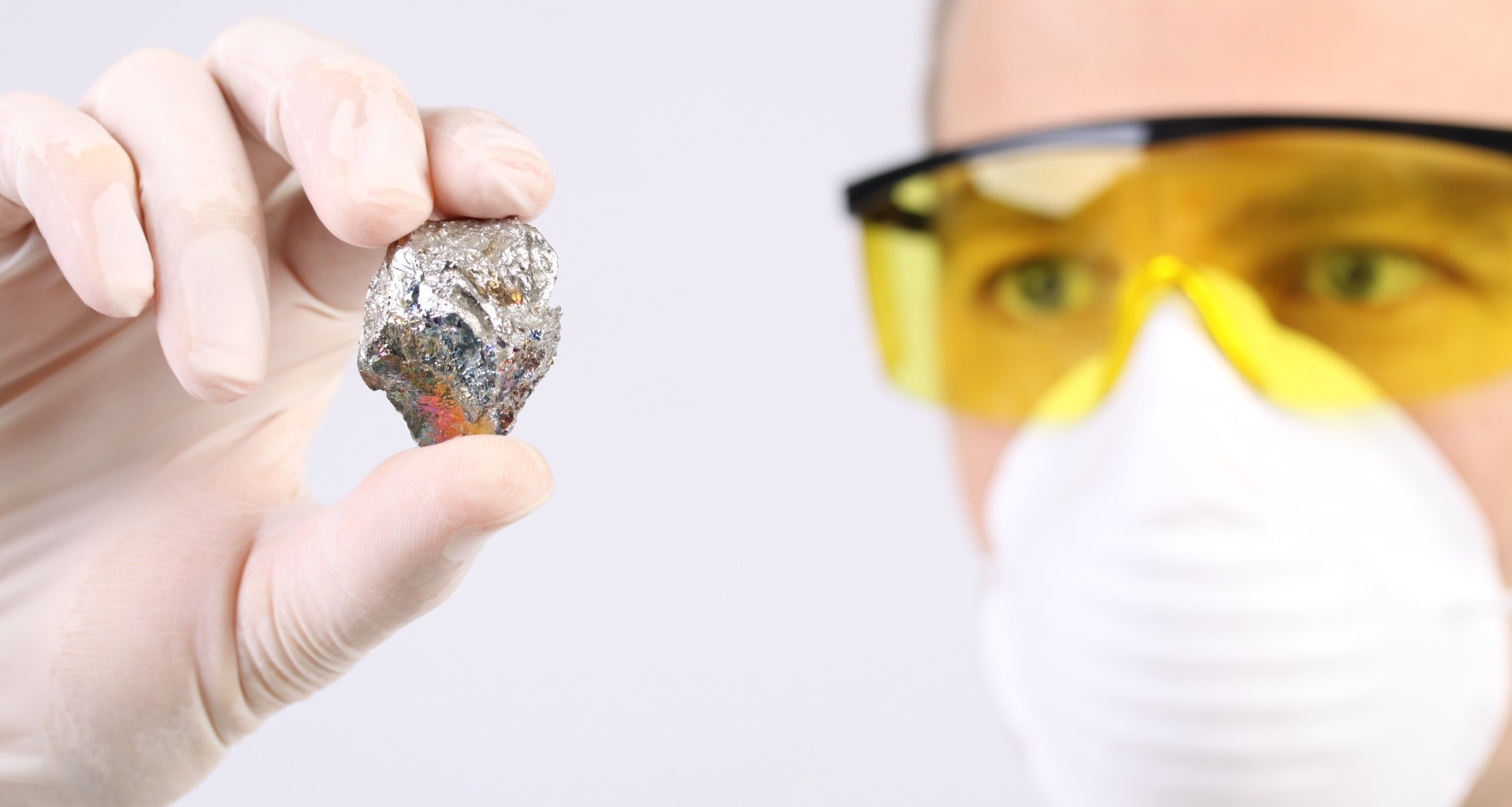 A scientist wearing yellow tinted safety specs holds a piece of metal in a white gloved hand.