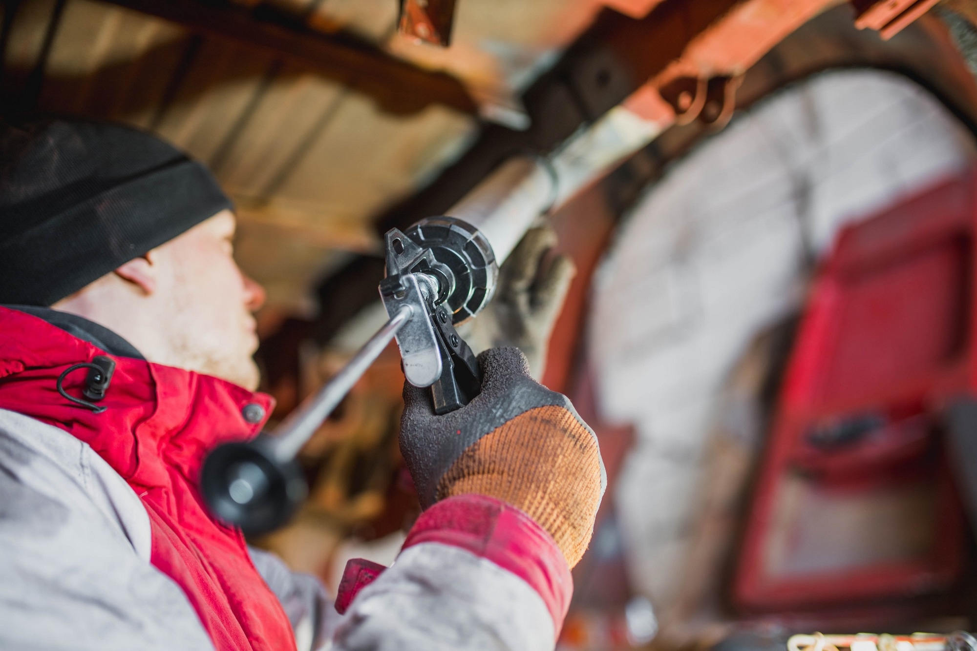 A car mechanic wearing a beanie and a red and white coat applies rust protection to the underside of a car.