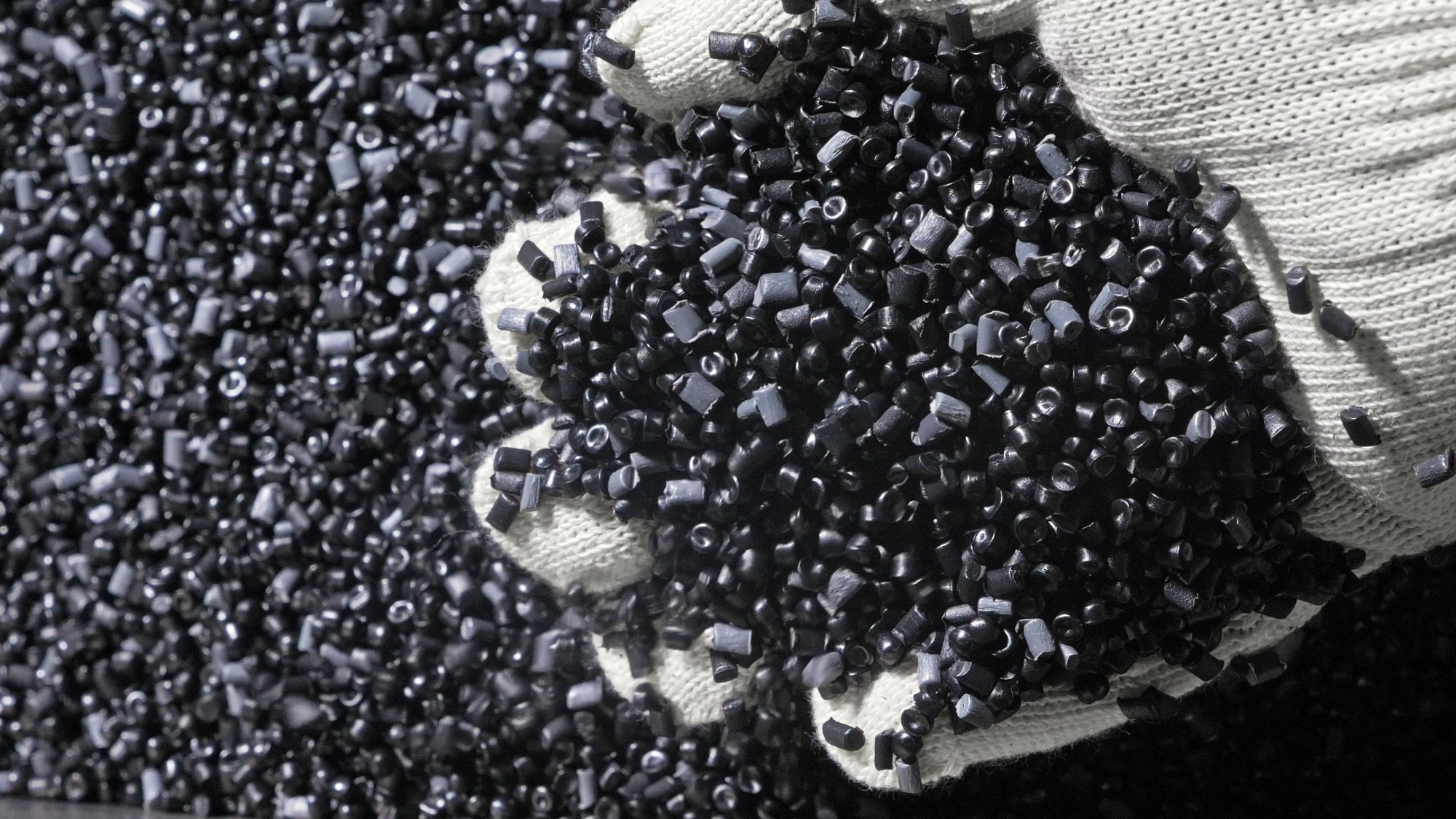 A gloved hand picks up black polymer beads from a vat of beads.