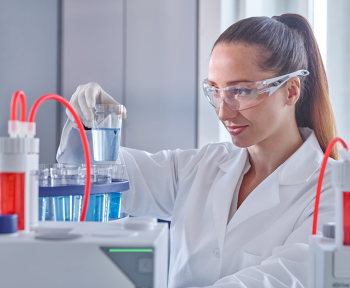Female scientists wearing goggles and gloves analyzing a beaker filled with lightly colored blue liquid.