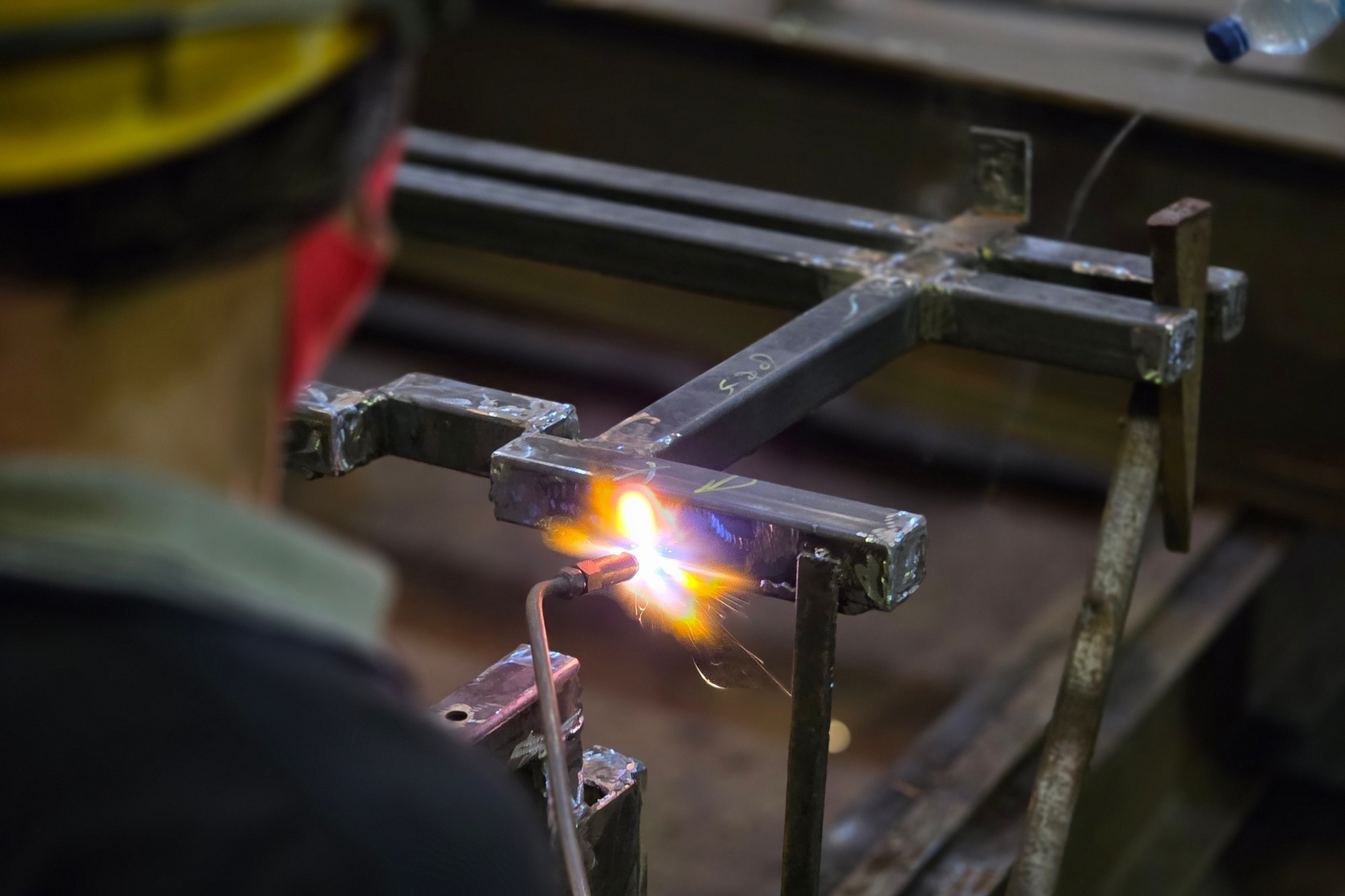 A man wearing a hardhat welds a corner of a piece of steel infrastructure.