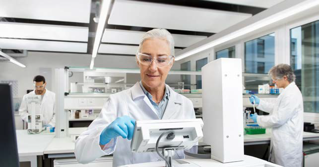 An older woman wearing spectacles and a lab coat adds a sample to a measurement instrument.