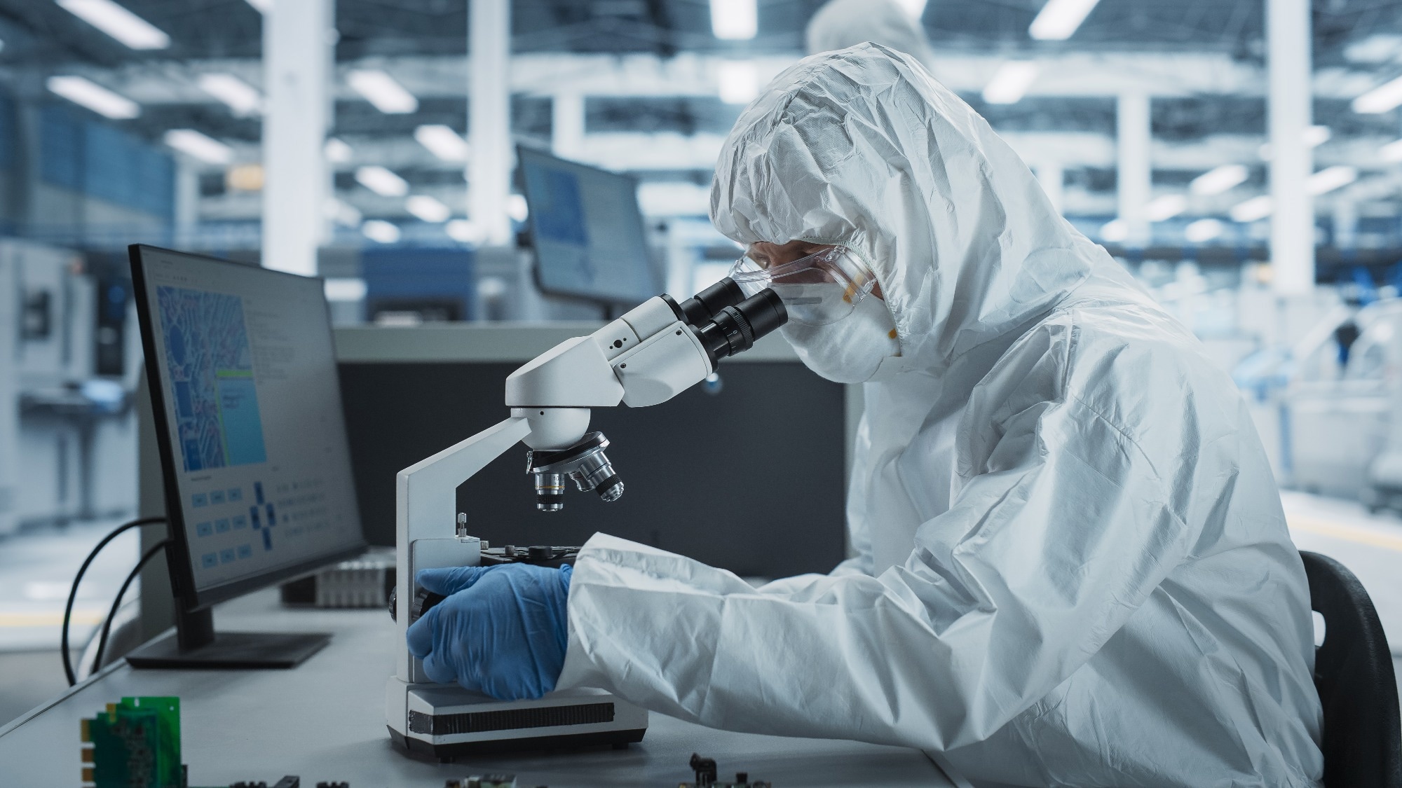 Two Scientists In Sterile Suites At Electronics Factory: Engineer Using Microscope And Desktop Computer To Inspect Microchip While His Colleague Using Soldering Jet Printer To Print Motherboards