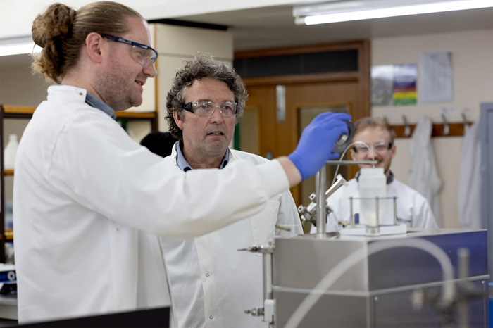 Two scientists (men in white lab coats with safety goggles) add a sample to an instrument in a lab.