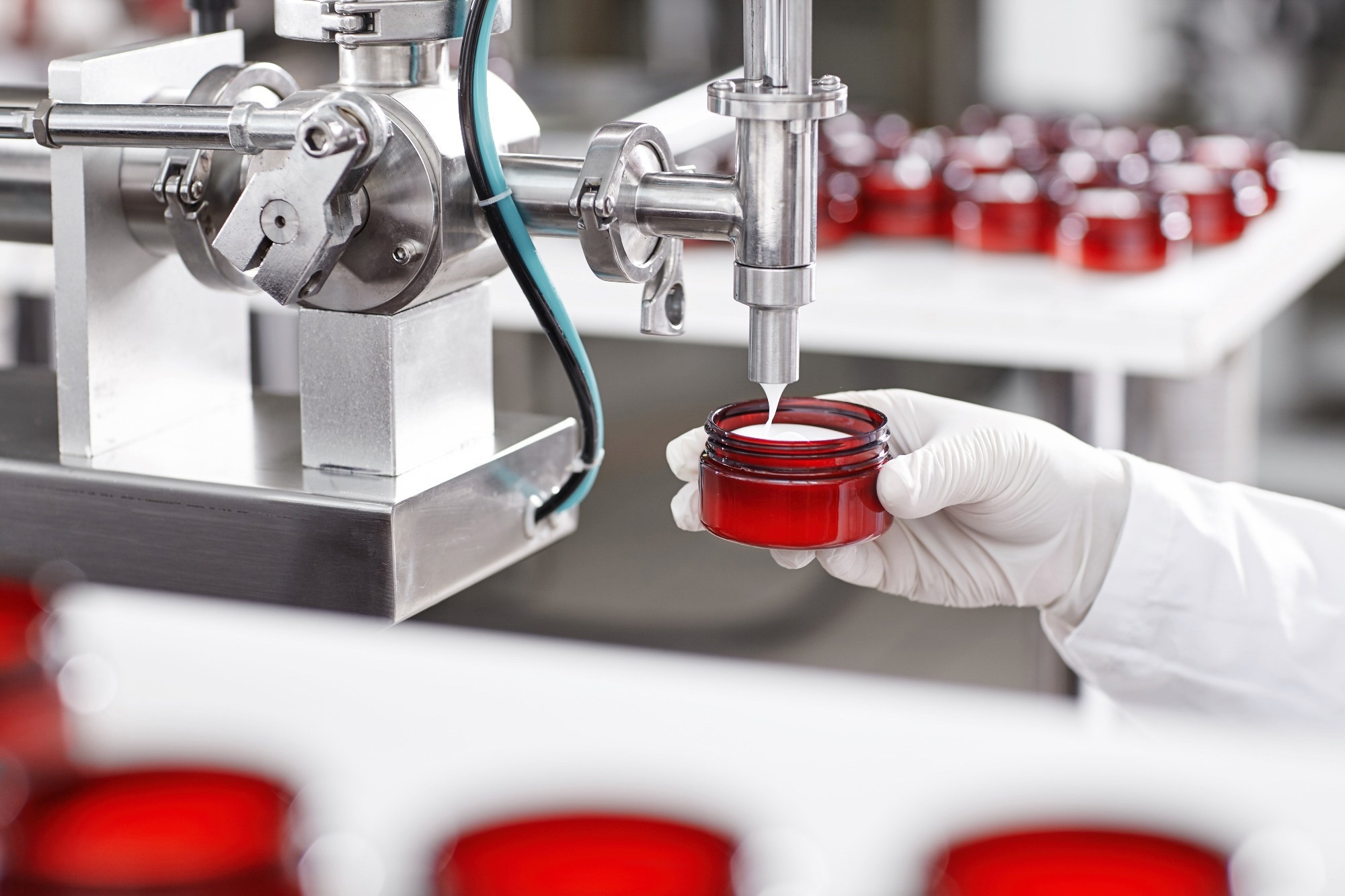 Researcher holding container under big metal tube pouring beauty cream isolated over working process at factory and many containers