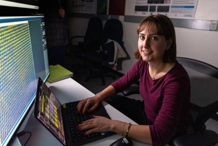 Female scientist smiling at camera, sat in front of a large PC