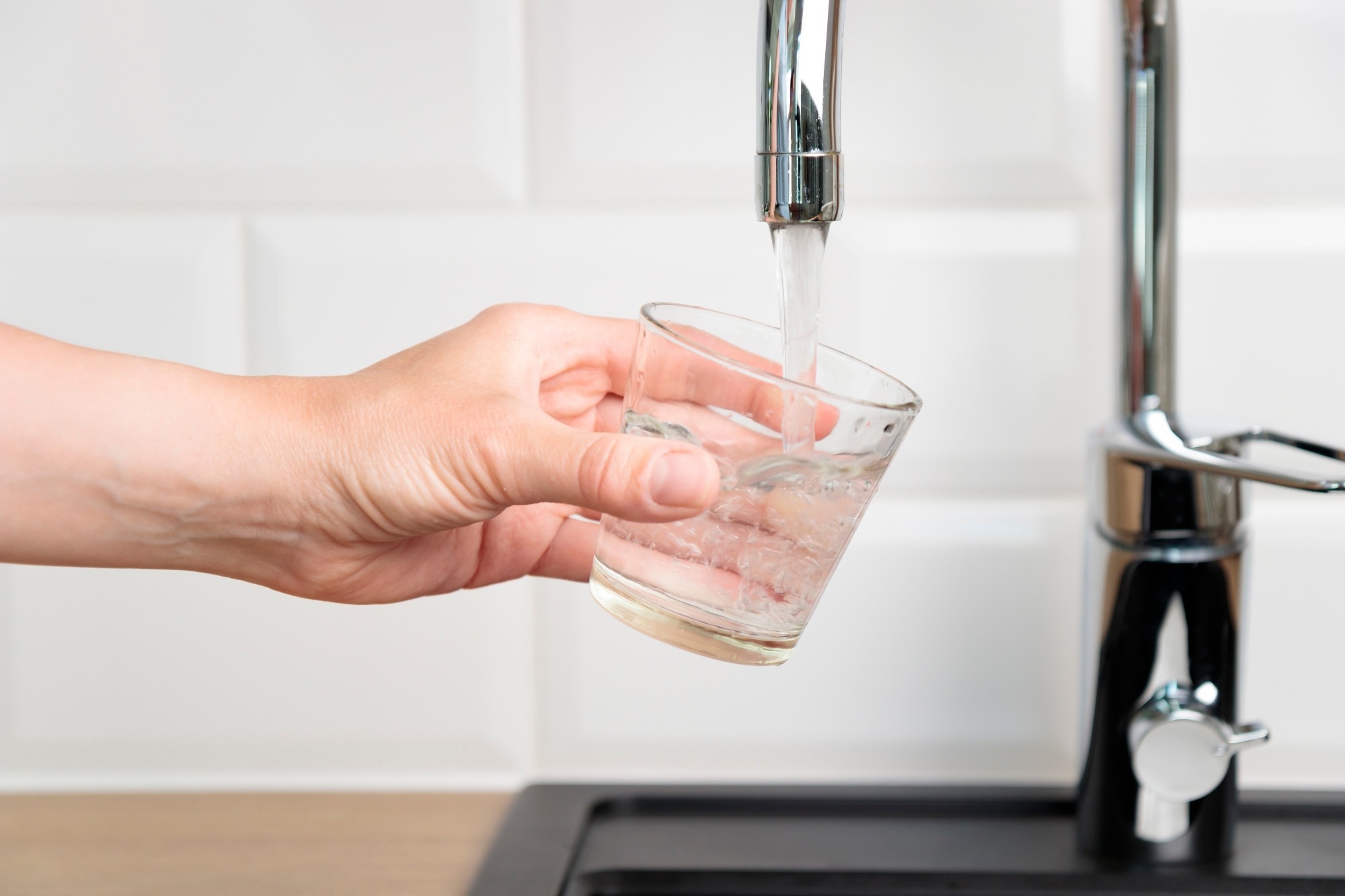 Person filling up a glass of tap pure water from kitchen faucet with white tiles wall.