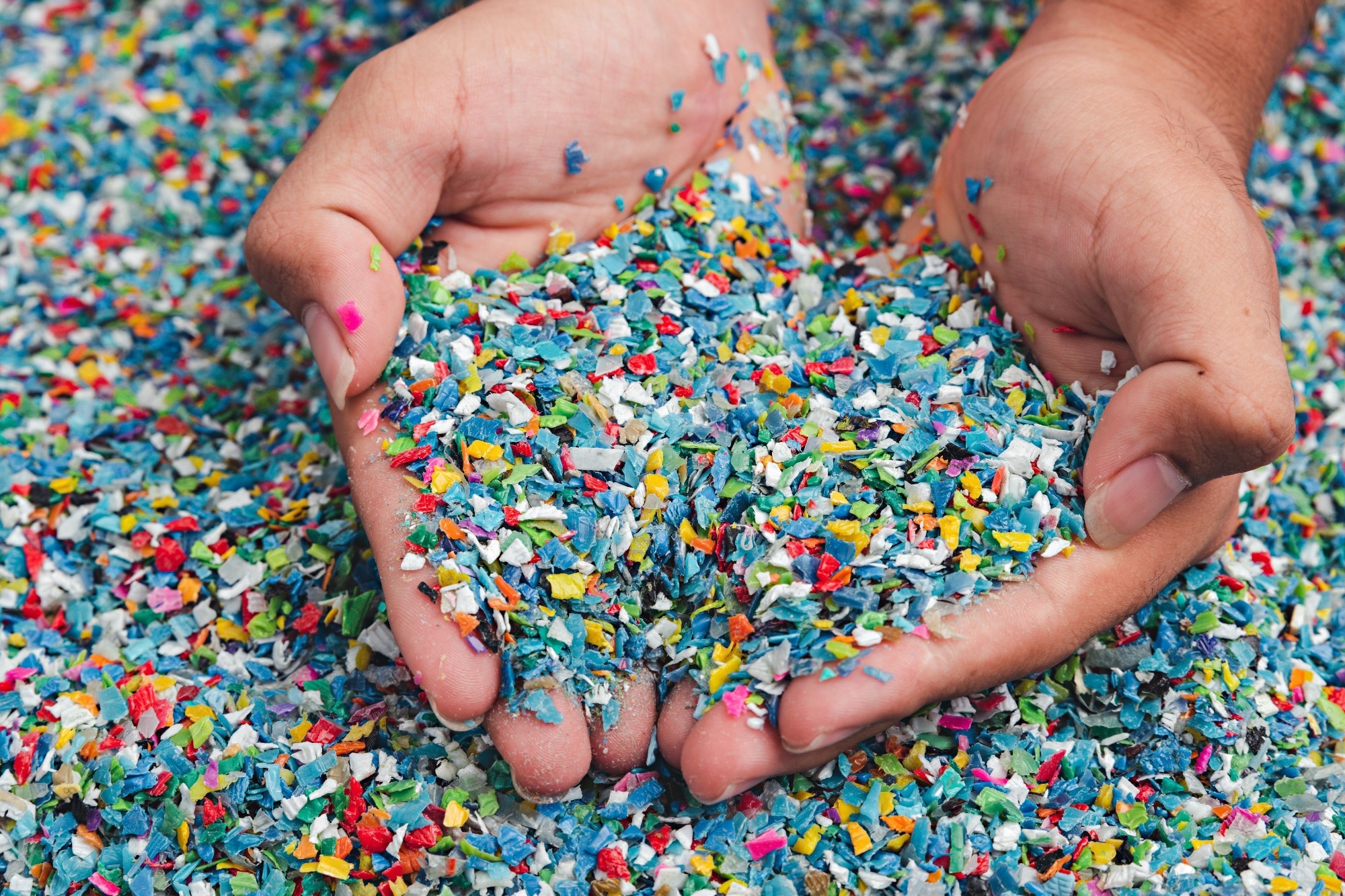 Close-up image of hands holding colorful shredded plastic pieces, with a large pile of mixed plastic waste in the background