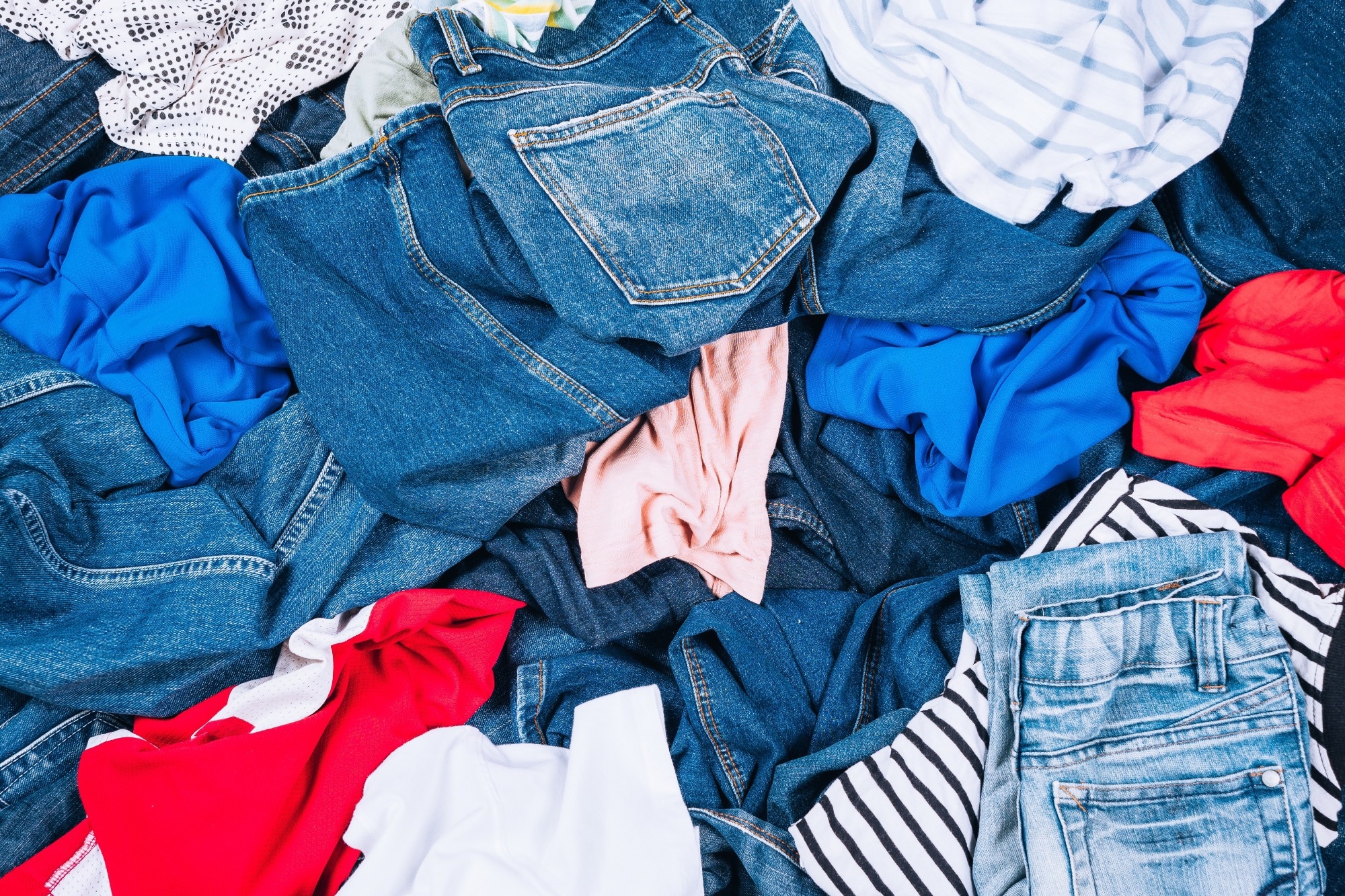 A pile of mixed used textiles including denim jeans, cotton shirts, and synthetic fabrics in various colors.