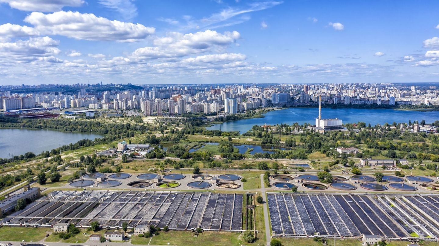 An aerial view of a city skyline with a lake, industrial facilities, and circular water treatment tanks surrounded by greenery.