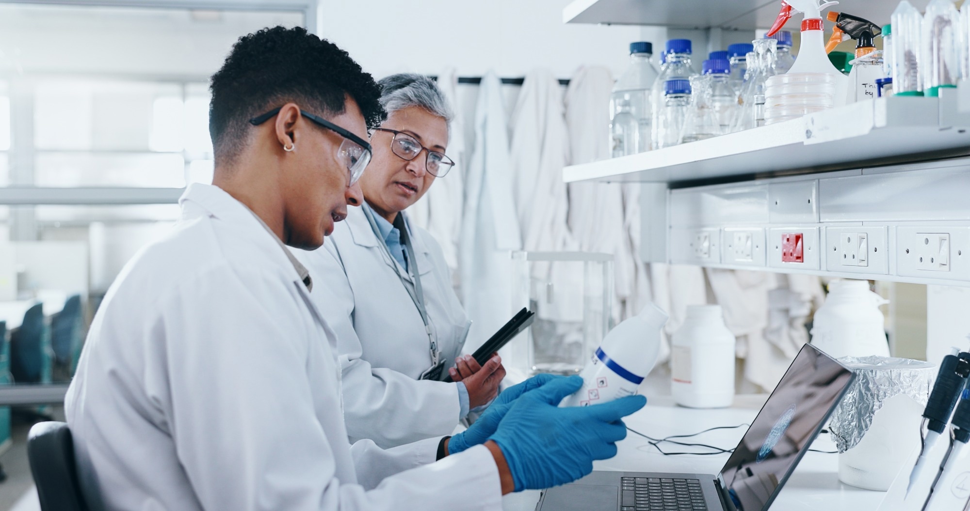 Two scientists examine a chemical label in a lab, wearing blue nitrile gloves.