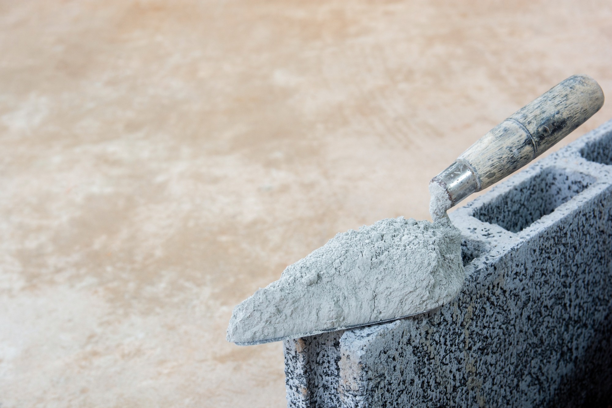 A cement laying tool (triangle spade) is placed on top of a breeze block in the foreground, with bare ground in the background.