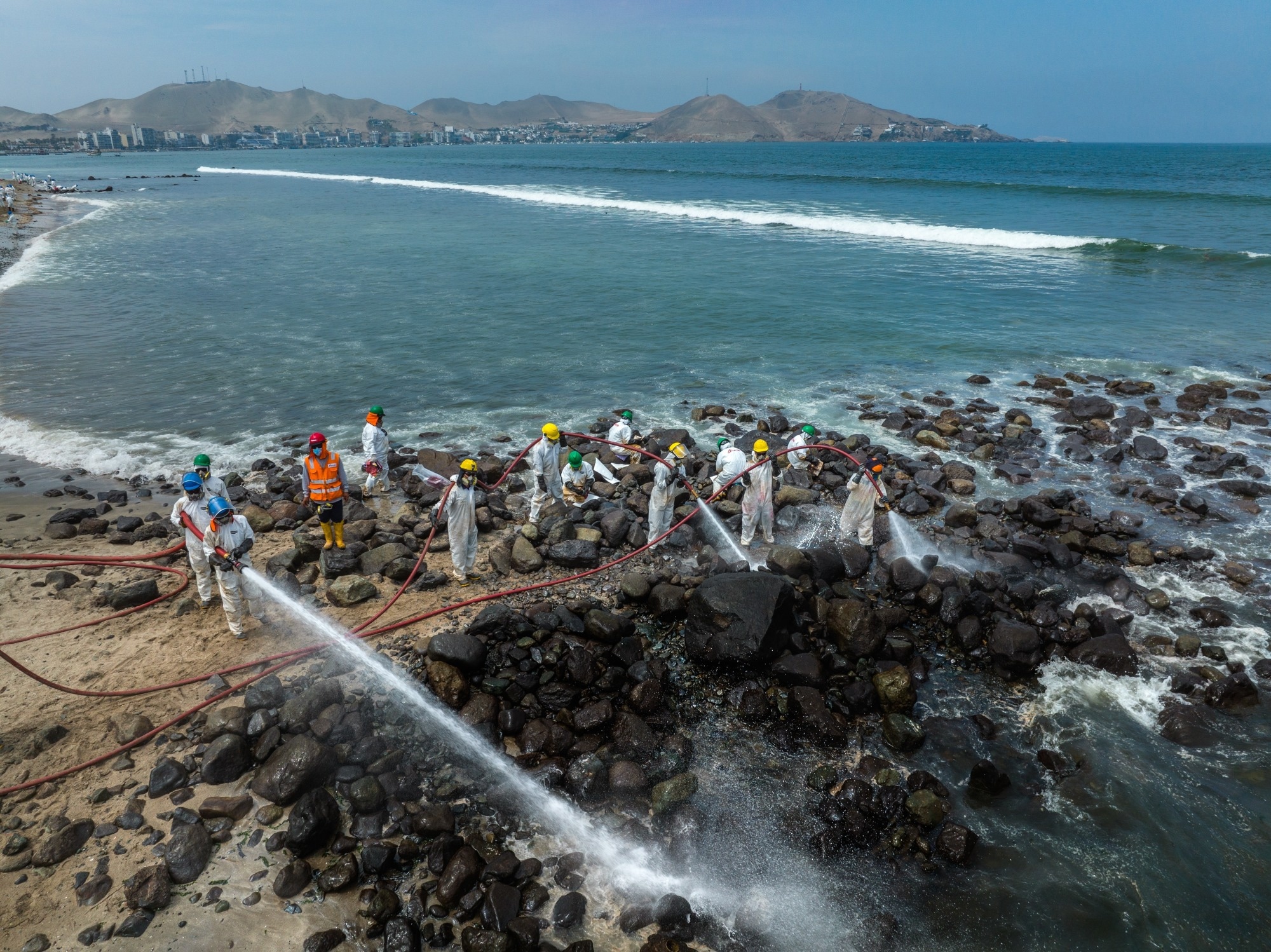 Aerial view of cleanup efforts after oil spill at "La Pampilla" refinery, Ancon, Lima, Peru on 2022. Workers in protective suits cleaning rocky shoreline.