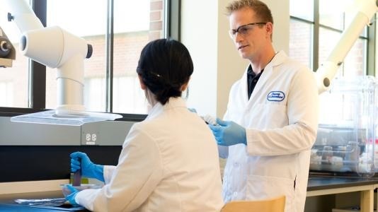 Jason Croy, director of the EaCAM consortium, talking to a fellow researcher. Both wear white lab coats and blue nitrile gloves.