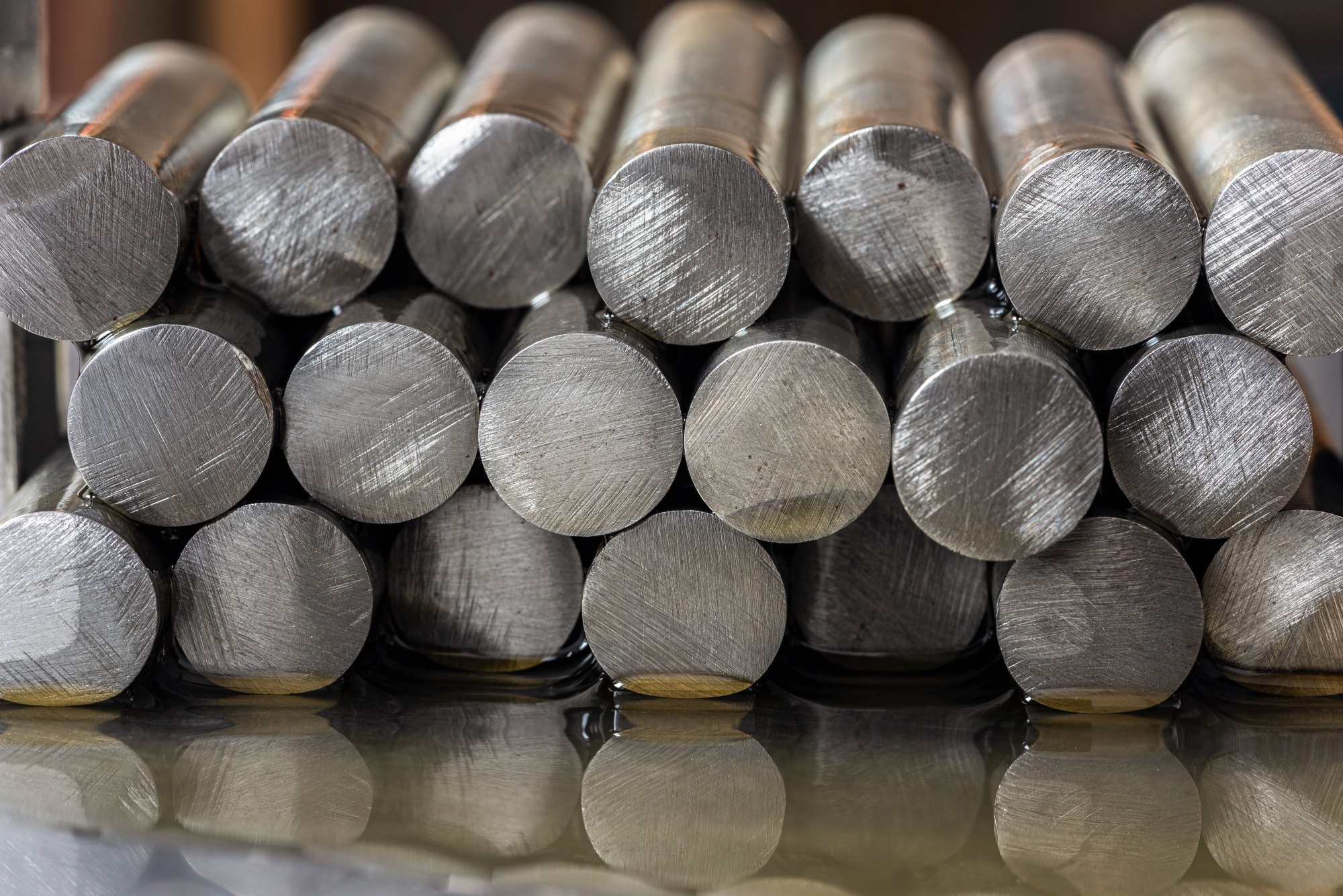 Stack of precious metal rods lying on an oil-covered metal surface with visible circular cross sections in industrial lighting.